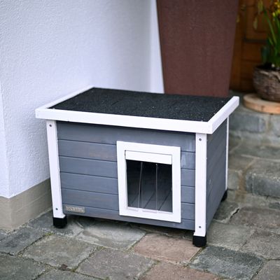 Серая деревянная будка для собак с белой рамой, черной крышей и прозрачной дверцей. Стоит на каменном полу у стены дома.