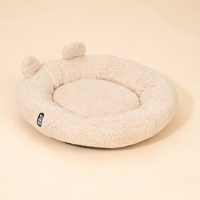 Rundes, beiges Liegekissen aus Plüsch mit erhöhtem Rand und zwei kleinen Ohren. Sichtbares Etikett mit der Aufschrift 'FGY'.