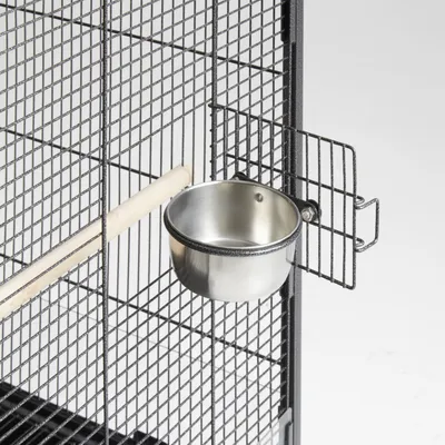 Gamelle en métal fixée à la porte d'une cage grillagée, avec un perchoir en bois visible à l'intérieur de la cage.