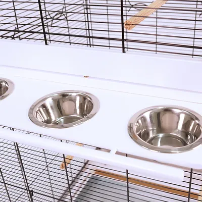 Trois gamelles rondes en acier inoxydable intégrées dans une cage à barreaux noirs, plateau blanc et perchoir en bois visible en arrière-plan.