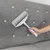 Hand holding a grey lint roller with handle, removing pet hair from a grey upholstered sofa.