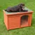 Animal lying on top of a wooden outdoor kennel with an arched entrance and plastic flap, shown on grass to demonstrate use and size.