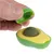 Jouet en forme d’avocat vert et jaune, avec une boule centrale marron, tenu par une main qui retire le couvercle. Inscription visible : TOYS Avocado.