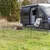 Mobiler Hundezaun auf einer Wiese neben einem schwarzen Camper. Ein Hund liegt innerhalb des Zauns, ein Stuhl mit Fellbezug steht daneben.