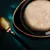 Close-up of a bowl with creamy food, visible chopped ingredients such as pieces of meat and vegetables. Next to it is a golden spoon on a dark background.