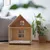 Wooden house with window design and soft insert, suitable as pet accommodation. Placed in a living room next to a plant.