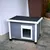 Grey wooden dog house with white frame, black roof and transparent door flap. Standing on stone floor in front of a house wall.