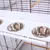 Three stainless steel feeding bowls in a white holder, mounted on a cage with metal grid and wooden perch.
