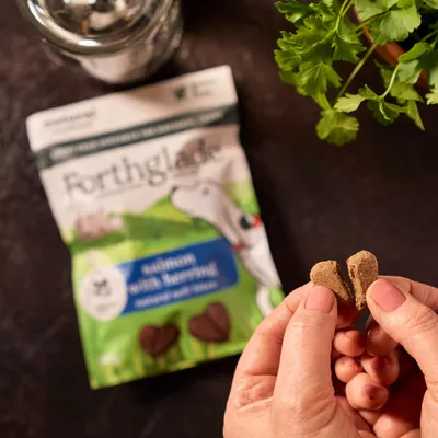 Hand breaking a heart-shaped treat in half, with a packet labelled 'salmon with herring' natural soft bites visible in the background.