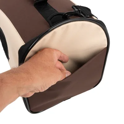 Main insérant un objet dans la poche extérieure d’un sac de transport pour animaux en tissu beige et marron, avec sangle noire visible.