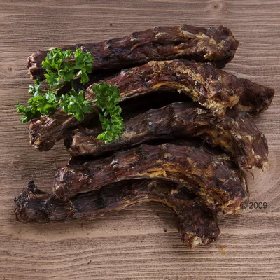 Несколько сушёных мясных шеек, уложенных на деревянной поверхности, сверху украшены веточкой петрушки. Видны текстура и цвет продукта.