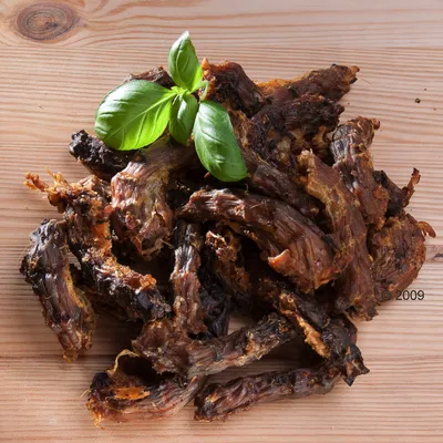 Кусочки сушёного мяса с зелёным листом базилика сверху на деревянной поверхности. Видны волокна и текстура продукта.