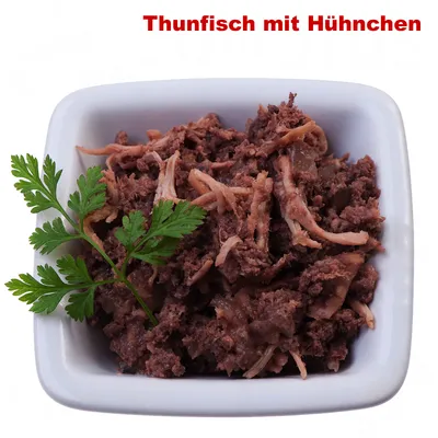 Texte en haut : Thunfisch mit Huhnchen. Mélange visible de thon et poulet effilochés dans une coupelle blanche, décoré d'une branche de persil.