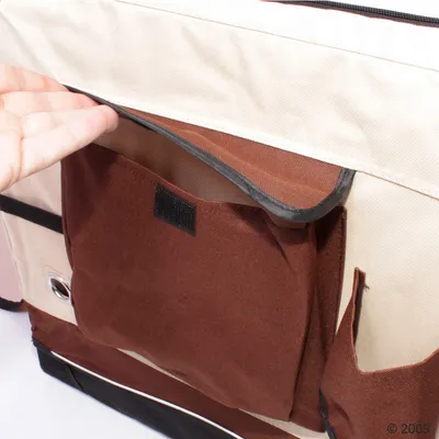 Close-up of a hand opening a brown fabric pocket with a Velcro fastening on the side of a beige and brown bag, showing storage compartment detail.