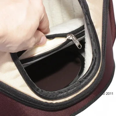 Detail van een hand die de ritssluiting opent van een beige en bordeauxrode draagtas met zachte witte binnenvoering, zichtbaar ritsvak en zwarte kunststof bodem.