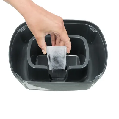 Mano colocando un filtro rectangular en el compartimento central de una fuente o bebedero de plástico gris oscuro para mascotas.