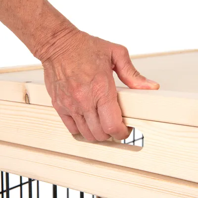 Main tenant une poignée découpée dans un couvercle en bois clair posé sur une structure à barreaux métalliques noirs, détail du système d’ouverture visible.