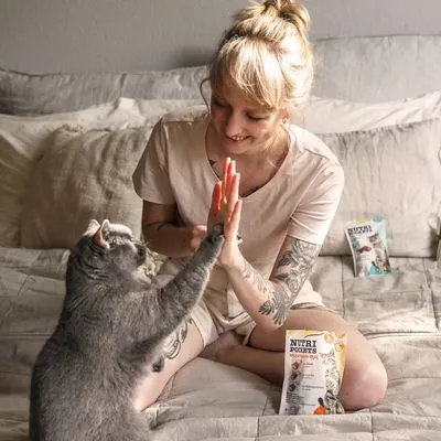 Grijze kat geeft poot aan persoon op bed, met twee zakjes Vitakraft Cat Nutri Pockets kattensnacks zichtbaar op de achtergrond en voorgrond.