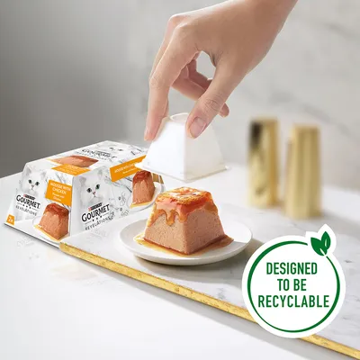 Gourmet Revelations Mousse with Chicken cat food, 2-pack packaging shown. Hand removing white plastic pot from mousse on plate. Badge reads: Designed to be recyclable.