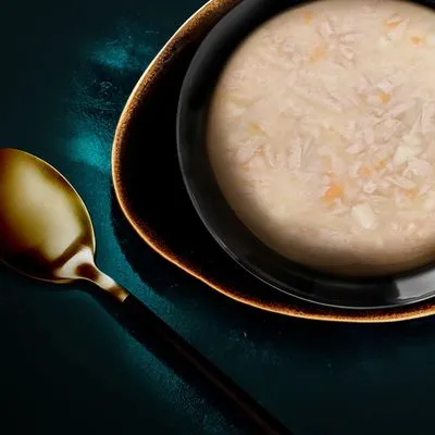Kom natvoer met stukjes in gelei, lichtbeige van kleur, geserveerd in een zwarte kom op een bord met een gouden lepel ernaast. Merknaam of productinformatie niet zichtbaar.