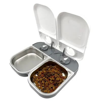 Automatisk dobbelt foderautomat med to åbne låg, to skåle i metal, én med tørfoder og én tom. Skålene er placeret side om side i en grå og hvid plastenhed.