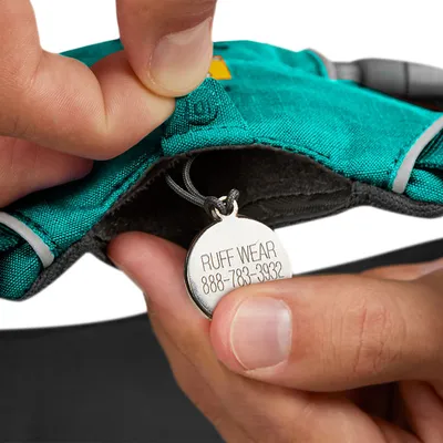 Medaglietta in metallo con testo visibile: RUFF WEAR 888-783-3932, attaccata a un collare o pettorina in tessuto verde tenuta da una mano.