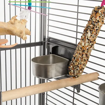 Intérieur de cage à oiseaux avec mangeoire en métal, perchoir en bois, jouet suspendu à perles colorées et bâtonnet de graines attaché aux barreaux.