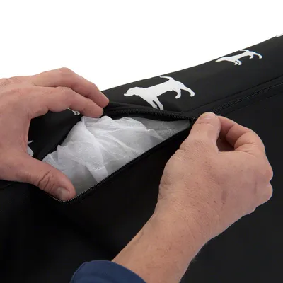 Detail rozepnutého zipu na černém textilním potahu s bílými siluetami psů, viditelná vnitřní výplň. Ruce otevírají zip.