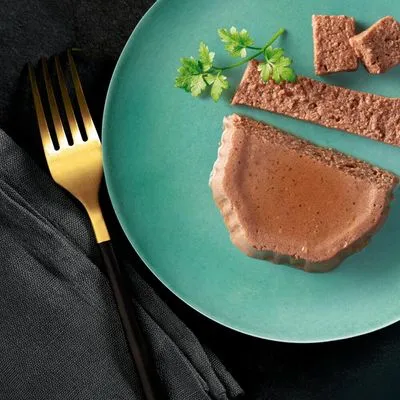 Portie natvoer in patévorm op groen bord, gesneden in plakjes en blokjes, met peterselie als garnering. Gouden vork en zwarte servet zichtbaar naast het bord.