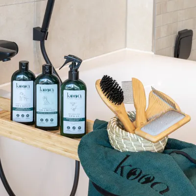 kooa Pflegeprodukte: Deep-Cleansing Shampoo, Sensitive Shampoo und Detangling Spray. Zubehör: Bürsten, Kamm und Handtuch mit kooa-Logo auf Bambusablage vor Badewanne.