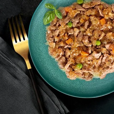 Natvoer met stukjes vlees, wortel en erwten in gelei op een groen bord, gegarneerd met basilicum. Gouden vork en zwarte servet zichtbaar naast het bord.