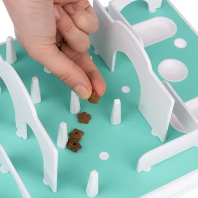 Hand places small brown treats on an interactive toy with white obstacles and a turquoise background.