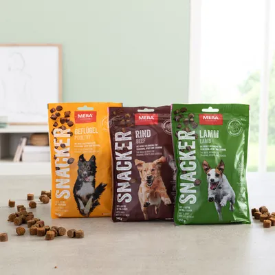 Trei pungi MERA SNACKER pentru câini: Geflügel Poultry (galben), Rind Beef (maro), Lamm Lamb (verde), cu imagini de câini și cuburi de recompense vizibile pe ambalaj.