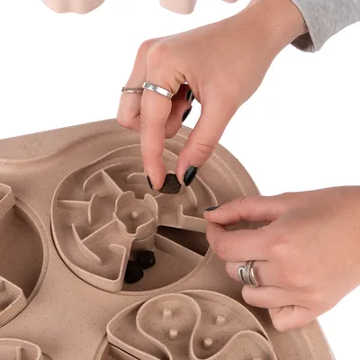 Händer placerar foderbitar i ett beige aktivitetsbräde med labyrintformade fack, tillverkat av plast. Flera foderbitar synliga i olika sektioner.
