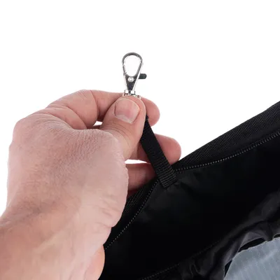 Main tenant une attache mousqueton en métal fixée à une sangle noire, partie d’un sac ou panier visible avec fermeture éclair noire.
