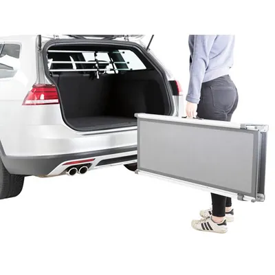 Osoba trzyma składany podjazd z metalu i plastiku przy otwartym bagażniku samochodu kombi. Wewnątrz auta widoczna kratka zabezpieczająca przestrzeń bagażową.