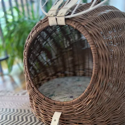 Pyöreä punottu kori, jossa on vaalea kukkakuvioitu pehmuste ja valkoiset nahkahihnat yläosassa. Materiaali: punottu rottinki, pehmuste kangasta.