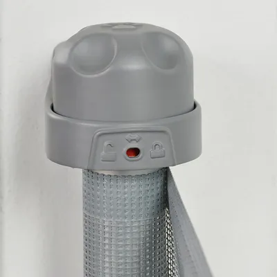 Bouton de verrouillage en plastique gris avec indicateur rouge, symboles de cadenas ouvert et fermé visibles sur la base, fixé à un filet grillagé gris.