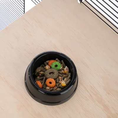 Black feeding bowl with mixed small animal food, consisting of grains, pellets and colourful rings on a light wooden table next to a cage.