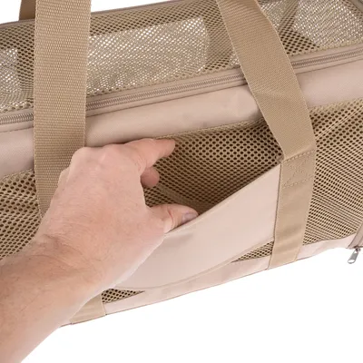 Sac de transport beige avec poche extérieure en maille, main ouvrant la poche. Poignées en tissu visibles, fermeture éclair sur le dessus.