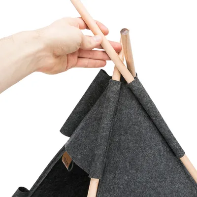 Mano che mostra il montaggio di un tipì in feltro grigio con pali in legno. Etichetta del marchio 'TRIXIE' visibile sul tessuto.