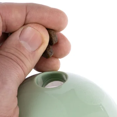 A hand fills brown treats into a green toy with an opening, suitable for keeping pets occupied.