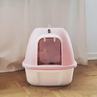 Затворена котешка тоалетна в розово с прозрачна врата и котка вътре. Марка Trixie видима отгоре.