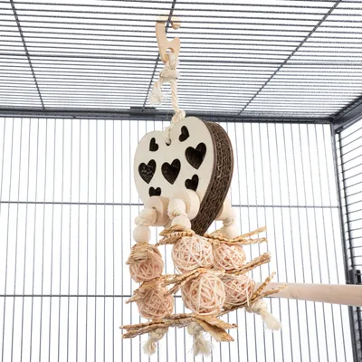 Jouet suspendu en forme de cœur avec découpes, boules et bâtonnets en bois et corde, accroché dans une cage pour petits animaux ou oiseaux. Matériaux visibles : bois, corde, rotin.