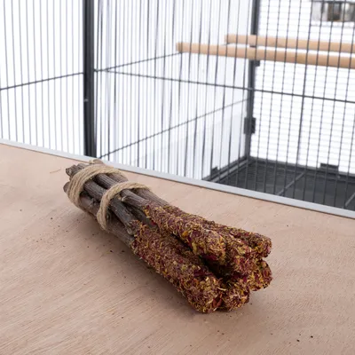 Összekötött fa pálcikák szárított virágokkal és gyógynövényekkel, egy faasztalon madárketrec előtt elhelyezve.