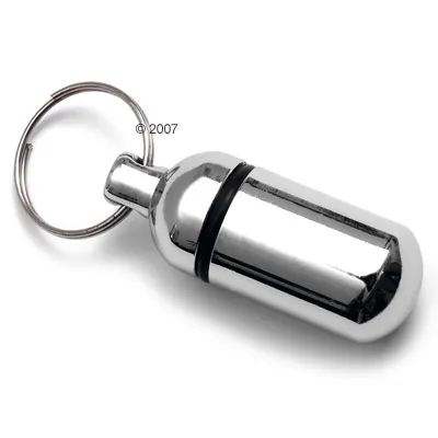 Capsule porte-adresse en métal argenté avec anneau porte-clés visible, joint noir au centre, sans texte apparent.