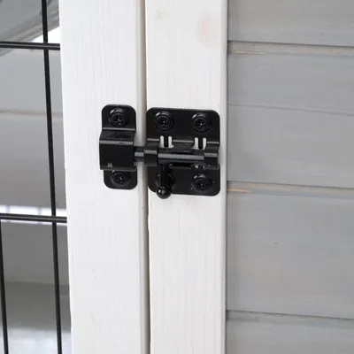Nahaufnahme eines schwarzen Metallverschlusses an einer weissen Holztür mit grauer Seitenwand und Gitterelementen.