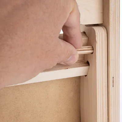 Hand showing how a wooden peg is inserted into an opening in a wooden frame.