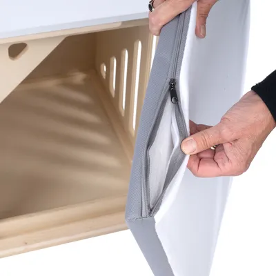 Hand öffnet Reissverschluss einer grauen Abdeckung, die einen Holzkäfig mit Lüftungsschlitzen verdeckt.