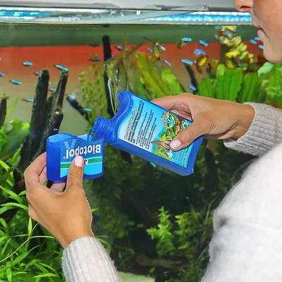 Person giesst Biotopol Wasseraufbereiter aus blauer Flasche in Messbecher vor Aquarium mit Pflanzen und Fischen.